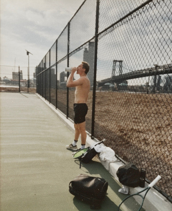 Man drinking from a bottle on a tennis court with tennis equipment and a cityscape in the background