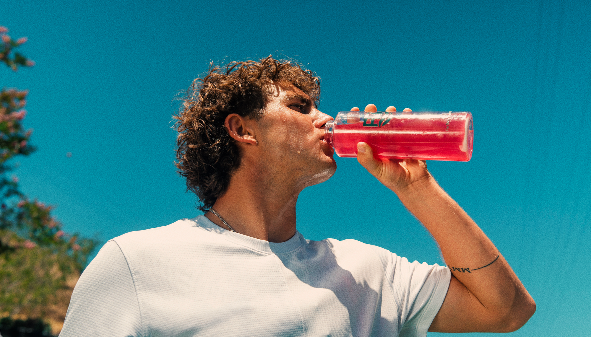 A man with curly brown hair drinking from a clear plastic water bottle filled with pink liquid, wearing a white t-shirt against a bright blue sky background.