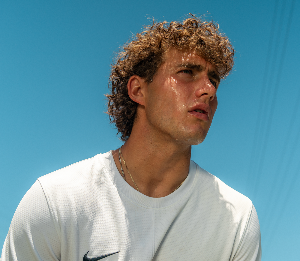 A close-up portrait of tennis player Tommy Holroyd wearing a white Nike athletic shirt against a bright blue sky background.