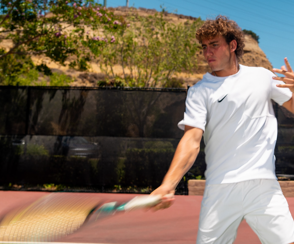 A male tennis player in a white Nike athletic outfit swings a racket on an outdoor clay tennis court, with a fence, greenery, and a hill in the background.