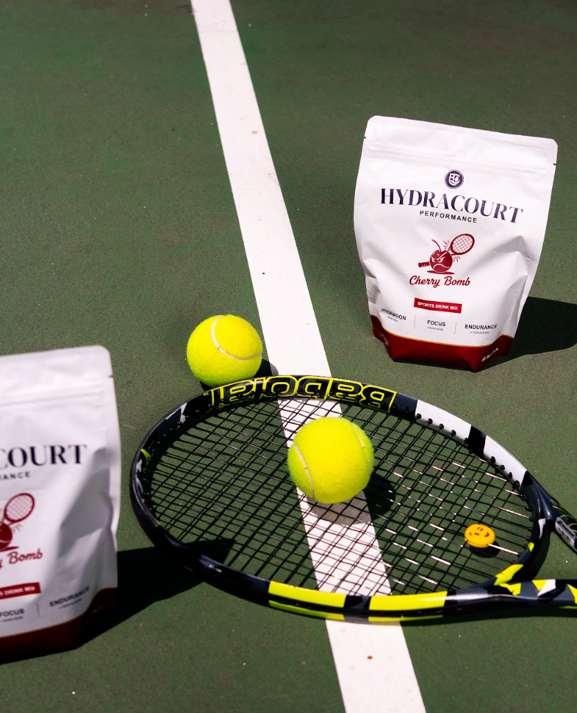 HydraCourt Cherry Bomb flavor tennis balls and product pouches arranged on a green tennis court with a racket and two tennis balls in the foreground.