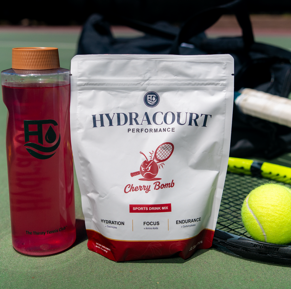 Hydracourt Performance Cherry Bomb sports drink mix pouch standing on a tennis court next to a pink hydration bottle, with a tennis racket and tennis ball in the background.